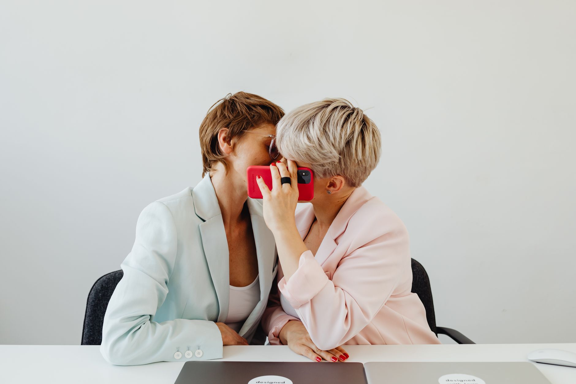 Two women share a secretive moment at a desk in a modern office setting.