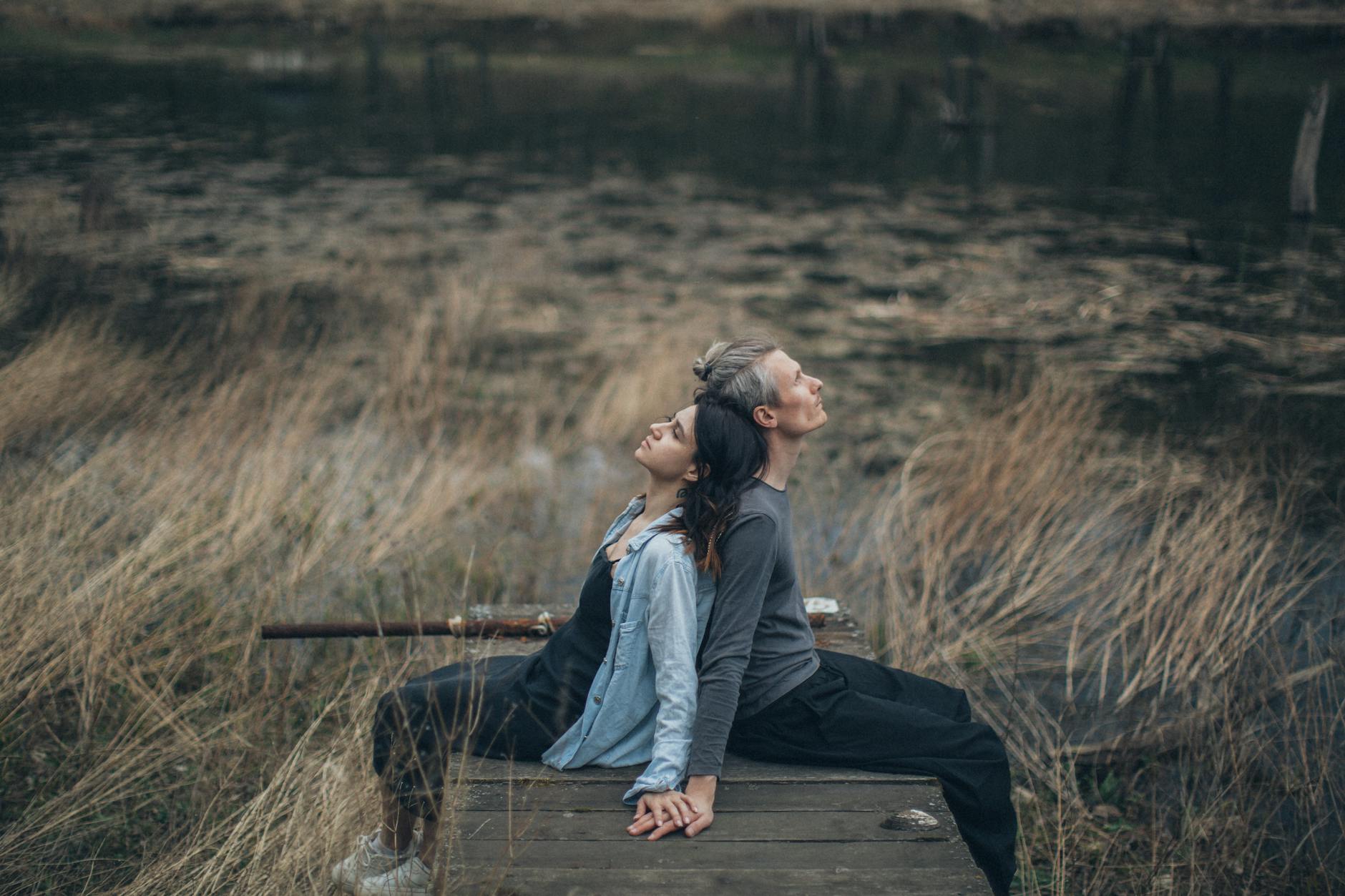Side view of content couple in casual clothes sitting together on wooden floor near pond and dry grass