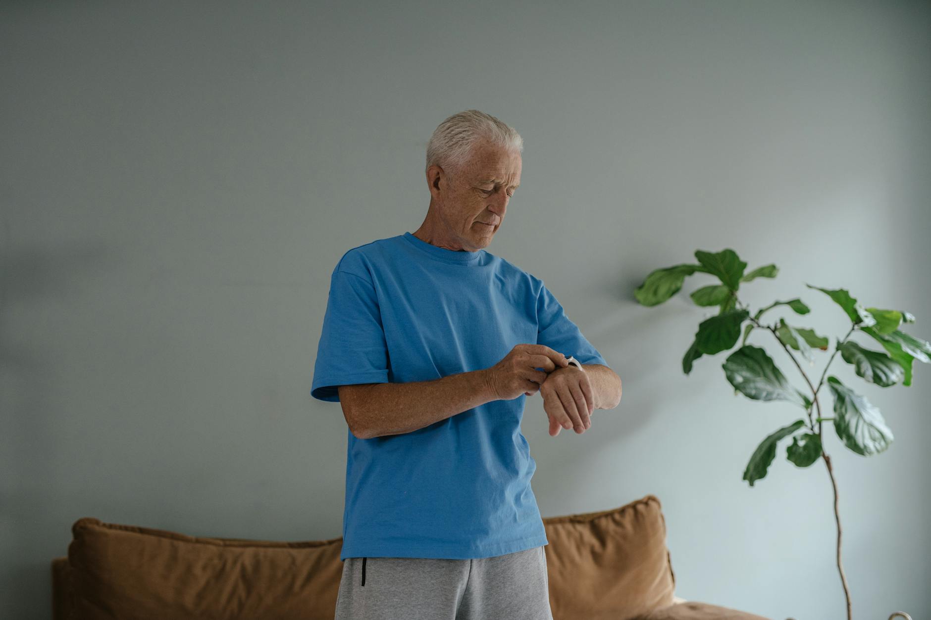 Elderly man in a blue shirt checking his wristwatch indoors by a plant.