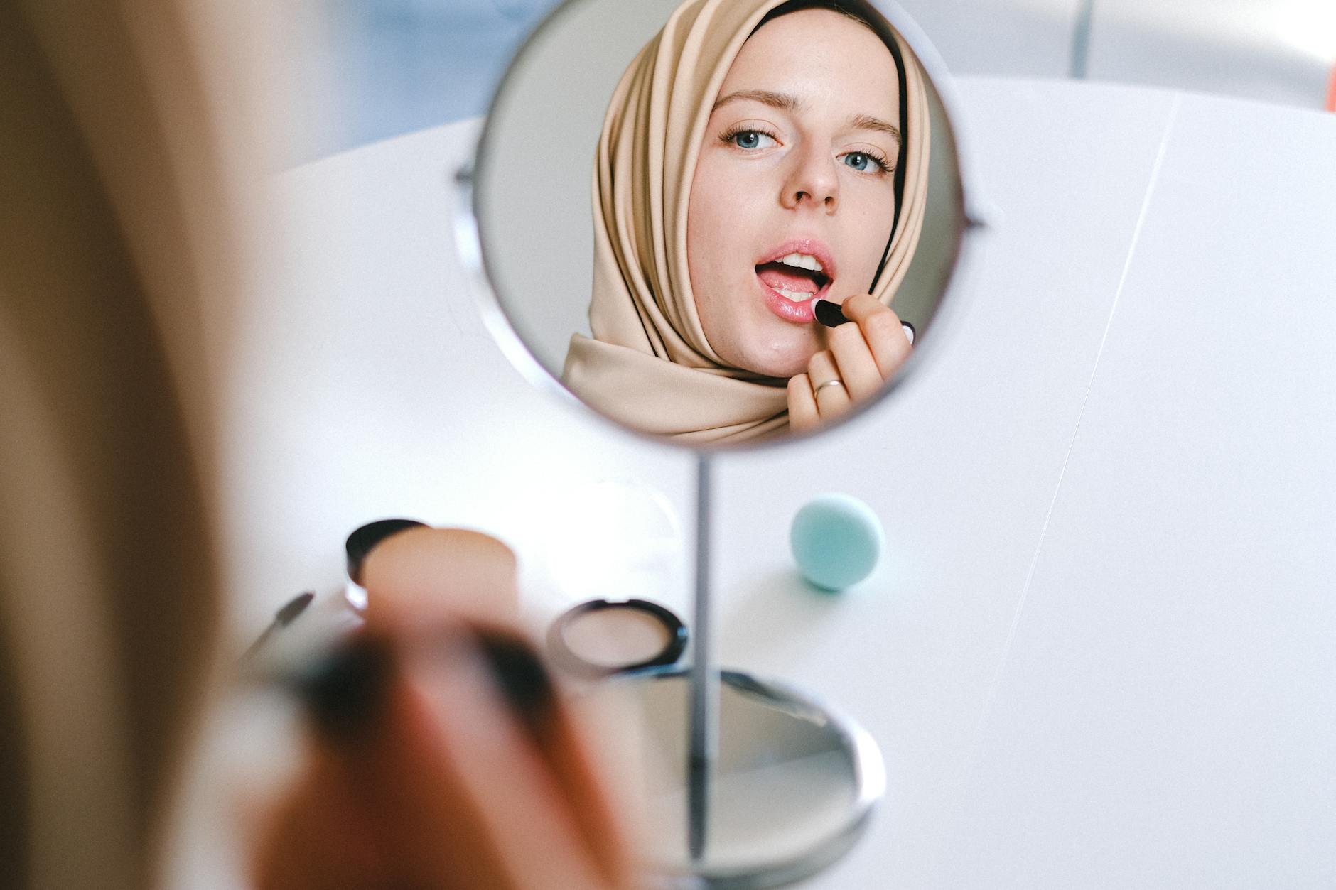 Muslim woman in hijab applies lipstick while looking at her reflection in a mirror.