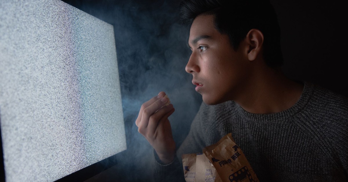 A young man engaged in watching TV static with popcorn in a dark room, creating a mysterious atmosphere.
