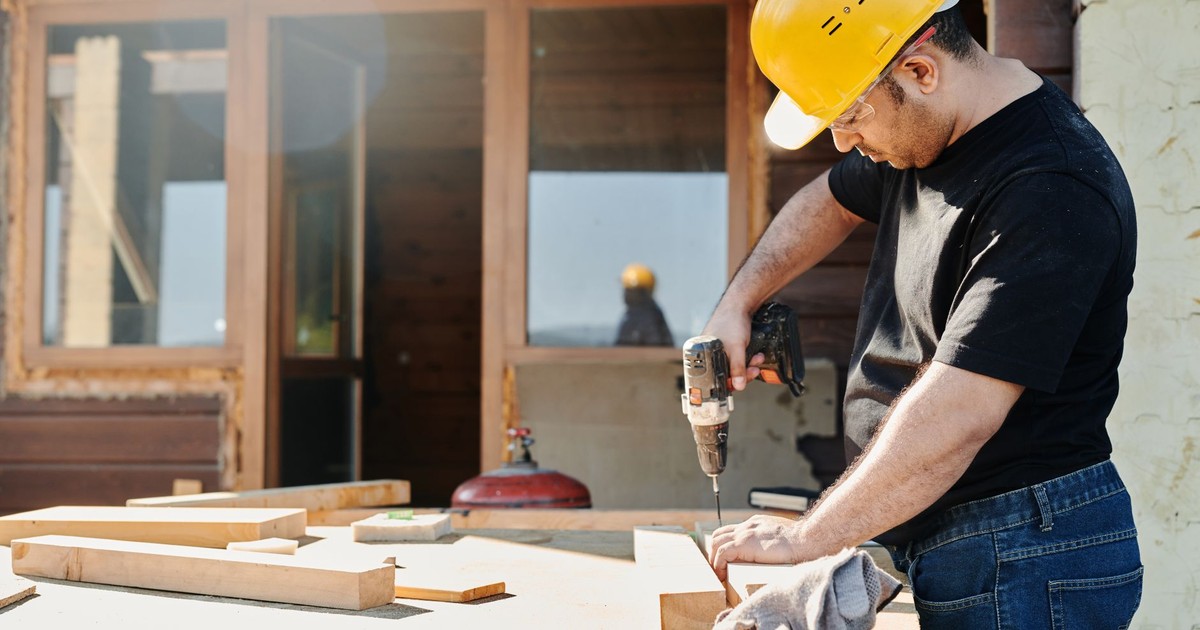 A construction worker with a hard hat uses a power drill on a wooden plank at a renovation site.