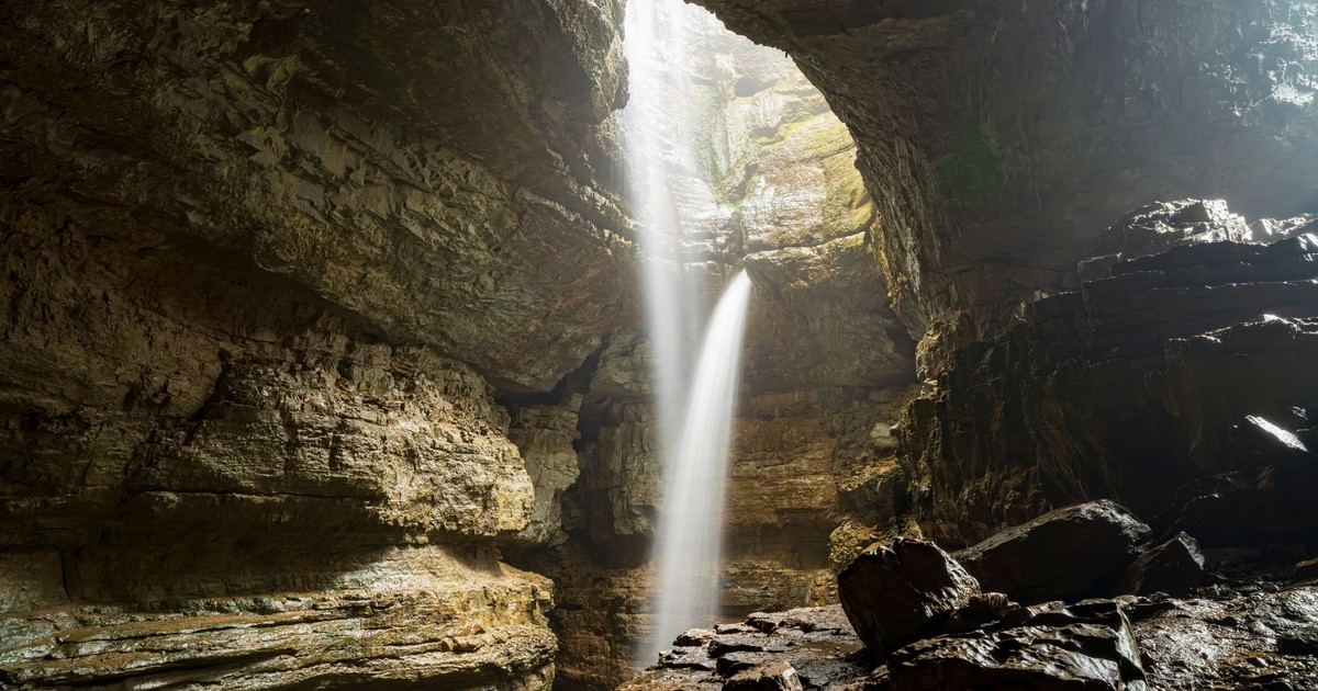 Sunlight illuminates a waterfall cascading in a tranquil rocky cave setting.