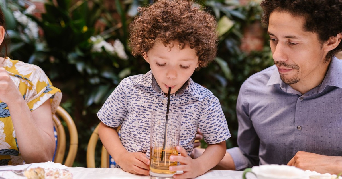 A child sipping a drink through a straw with father at an outdoor café table.