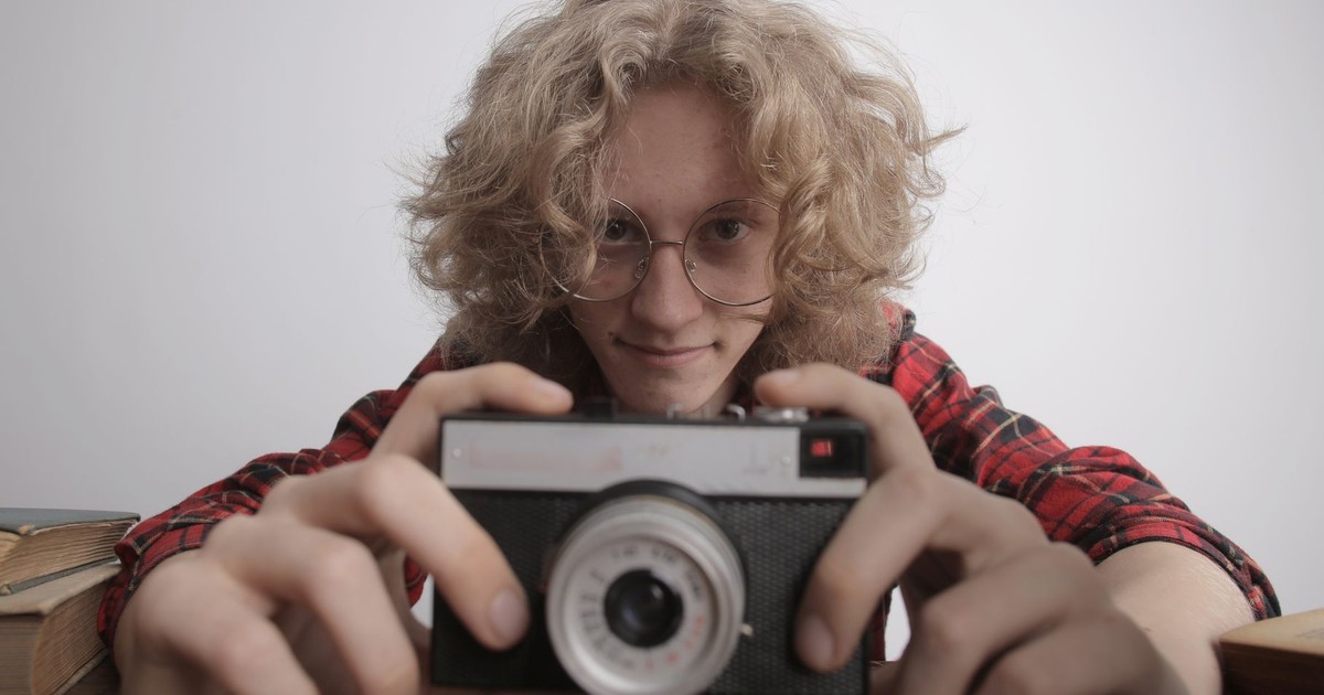Pleased teenage guy in red casual shirt and eyeglasses with curly long hair sitting at table and looking at camera while holding film camera in hand