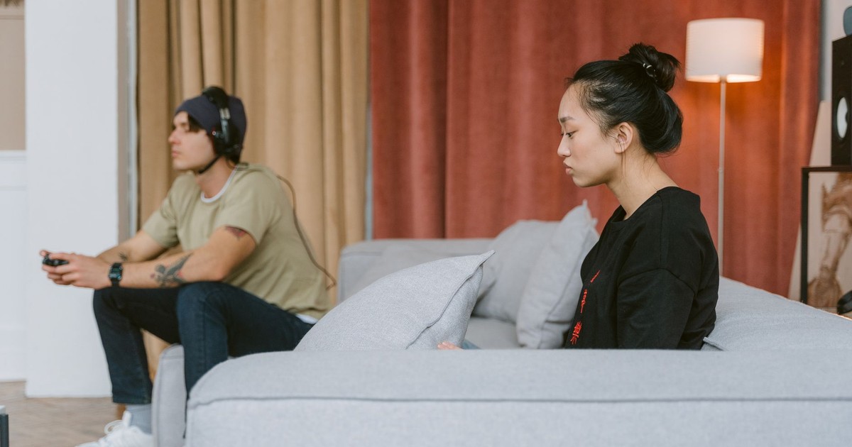 A couple sits on a couch, illustrating tension as one plays video games and the other appears distant.