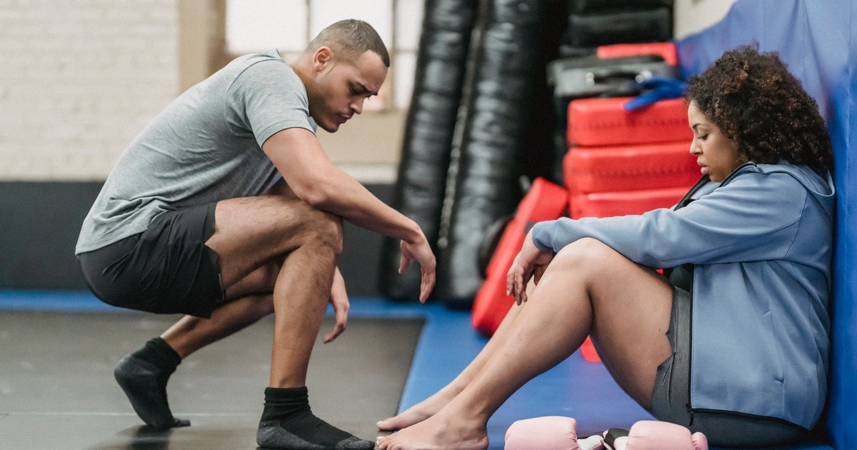 Side view full body of muscular black personal trainer sitting on haunches near tired African American female in gym with equipment