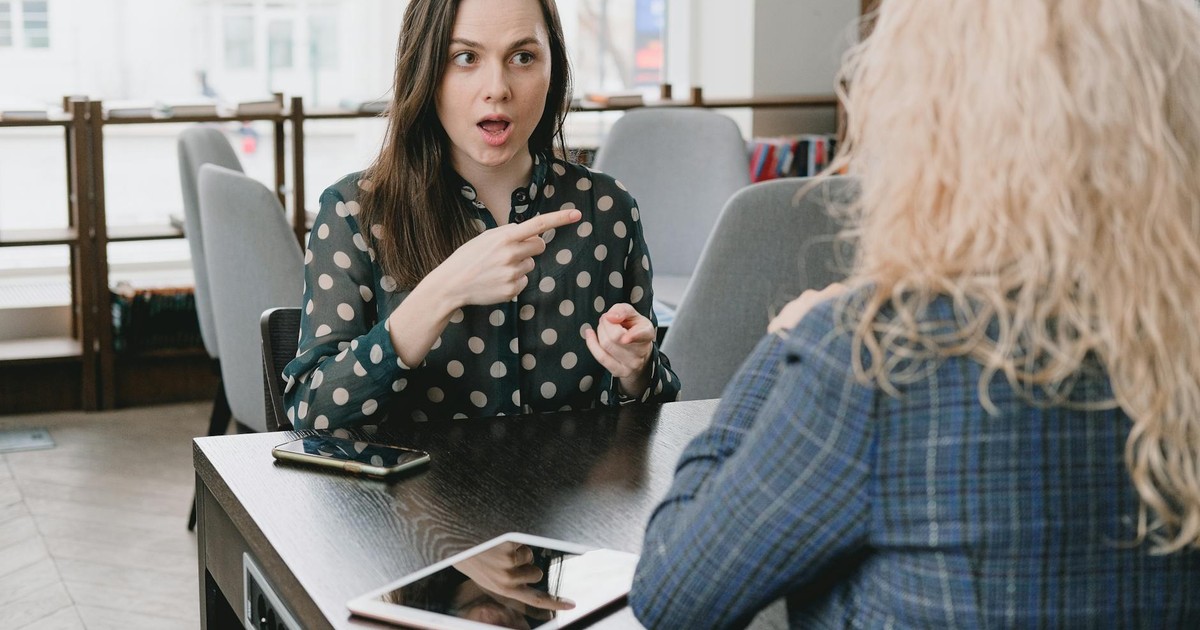 Astonished female pointing away while sitting at table with smartphone and tablet and talking to woman