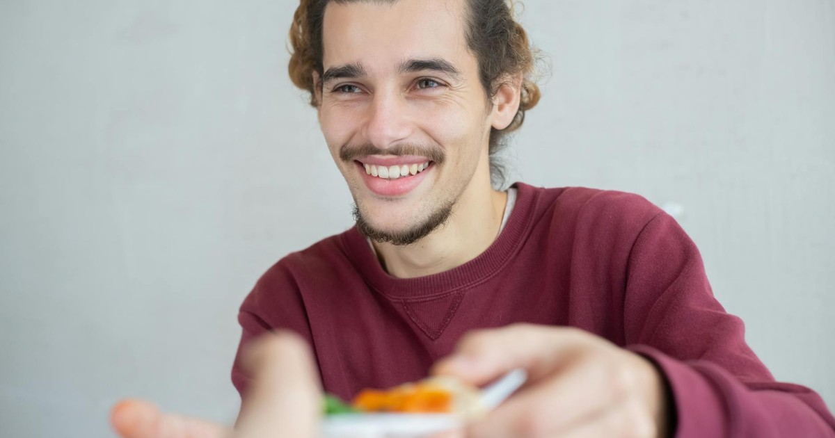Smiling young man with beard and mustache sharing a meal indoors, expressing warmth and friendliness.