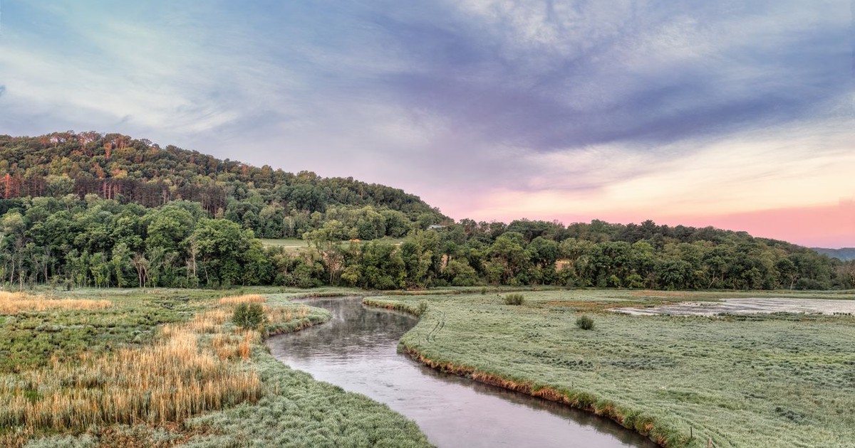 A tranquil river meanders through a lush valley at dusk under a dramatic sky.