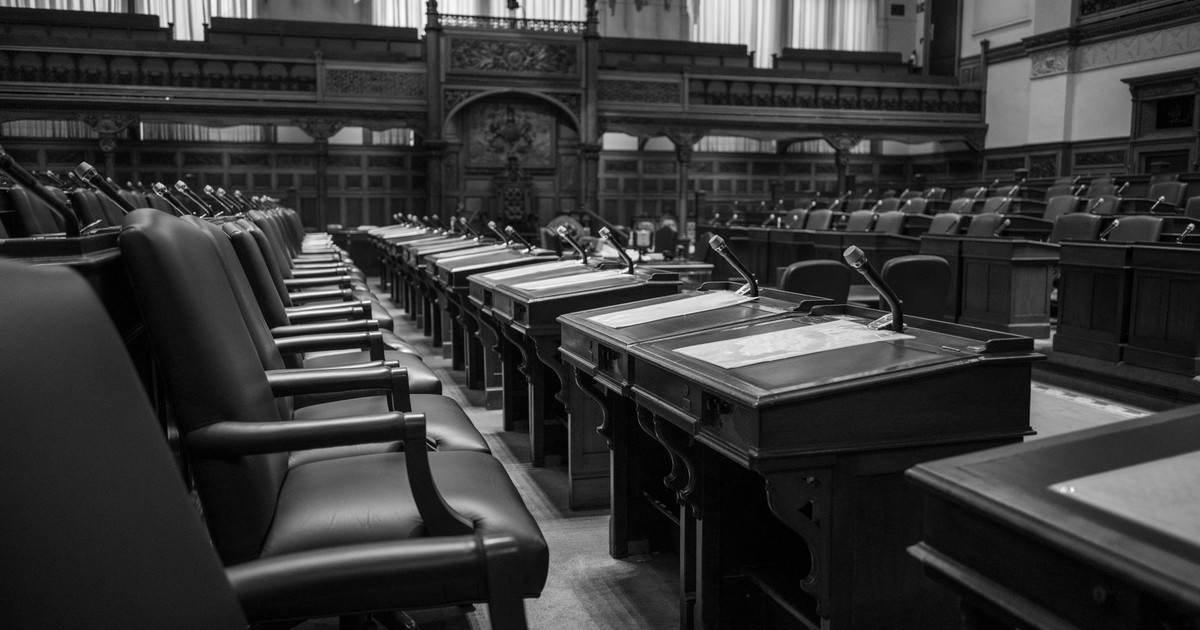 Black and white photo of a traditional legislative assembly chamber, featuring empty seats and desks.
