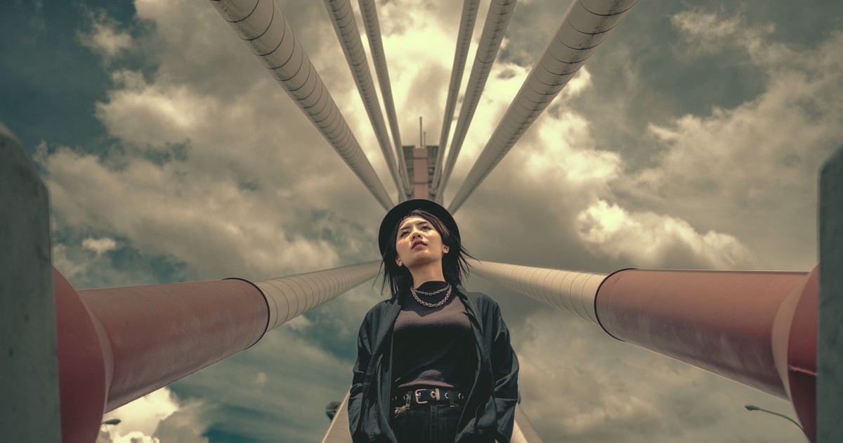 From below of young stylish female in casual outfit and hat standing near metallic red and white pillars with hands in pockets against cloudy sky