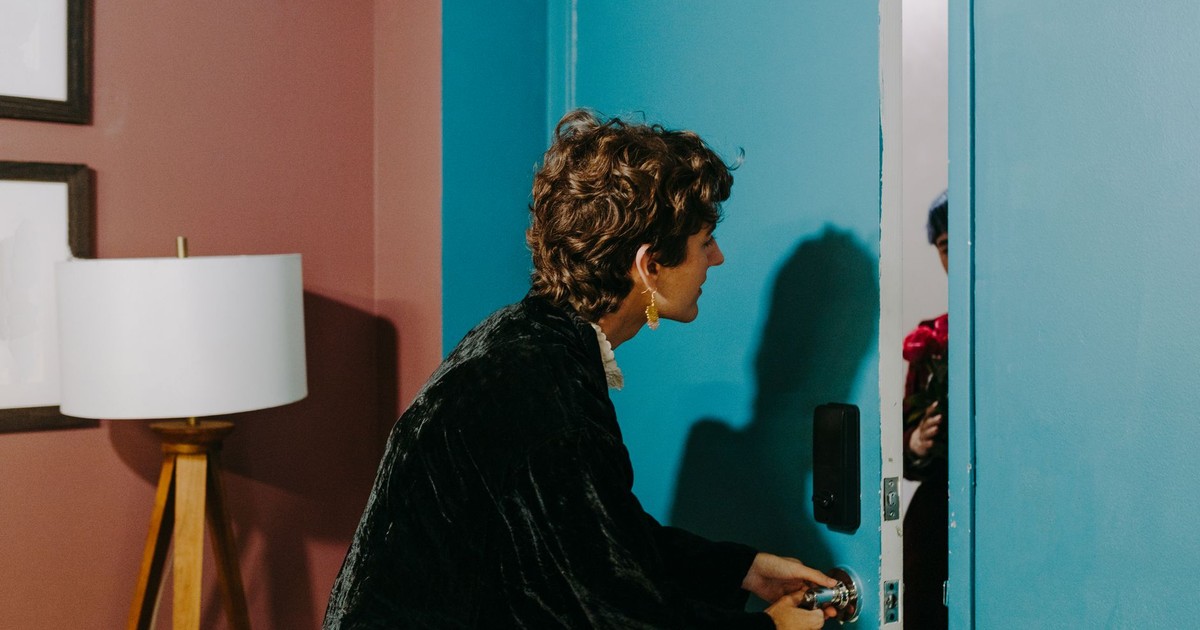A person at a blue door surprised by a visitor holding flowers indoors.