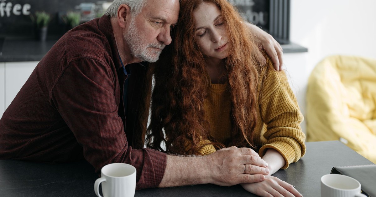 An elderly man consoles a young woman at a table in a warm, intimate indoor setting.