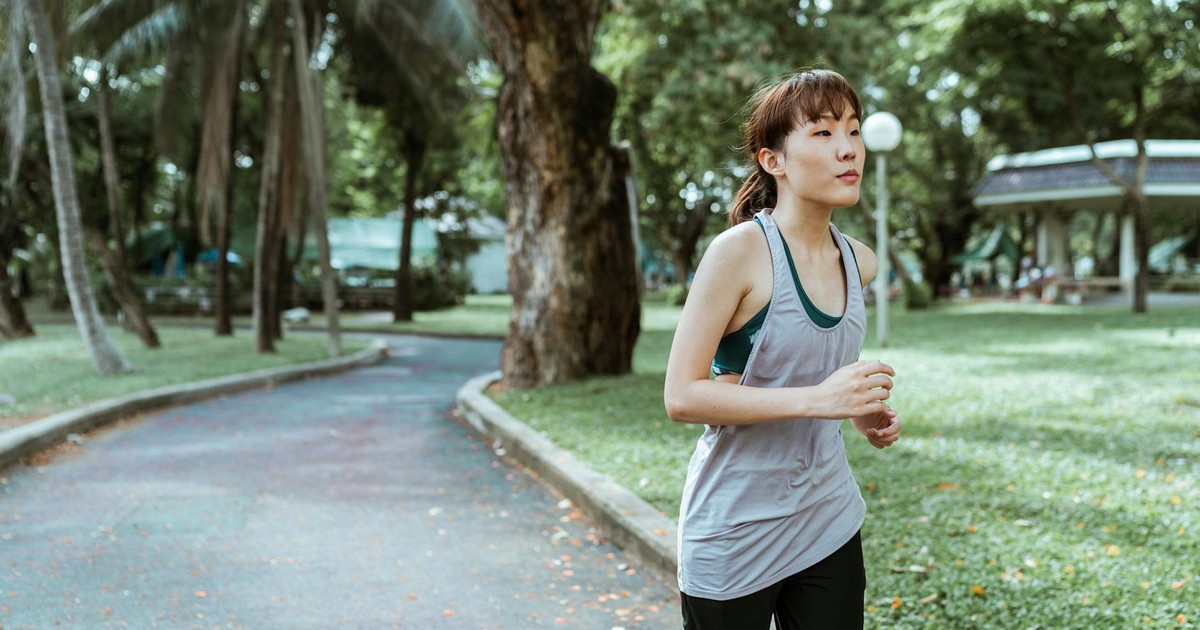 Thoughtful young Asian female athlete in sportswear running on asphalt road covered with faded leaves in city park in daytime and looking away
