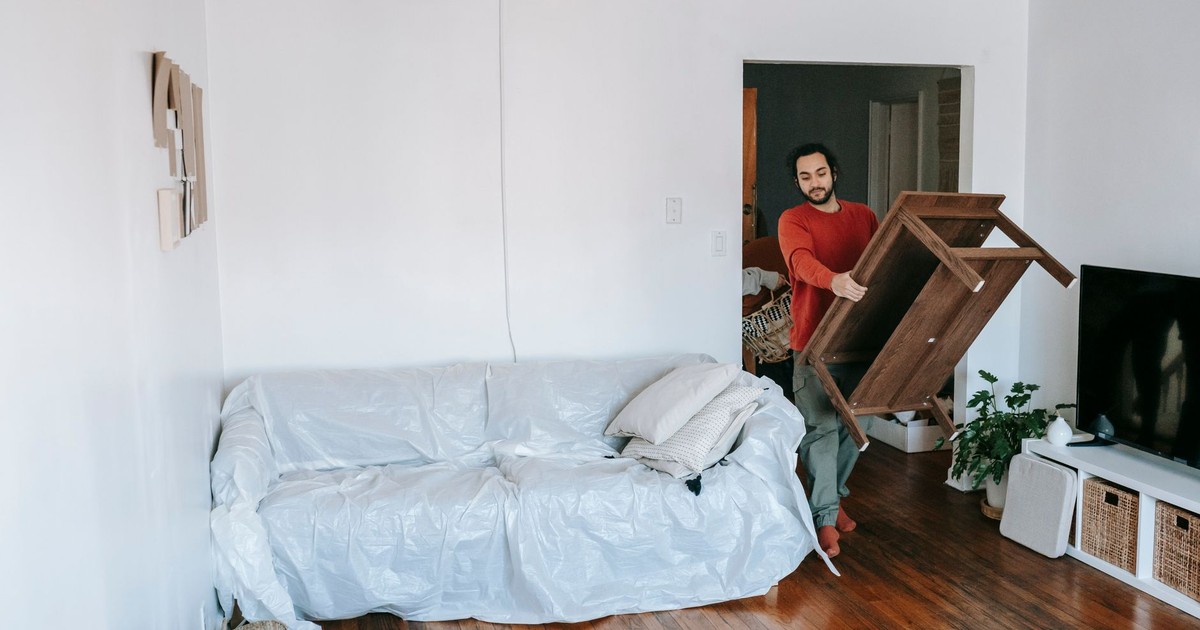 Man lifting a wooden table in a bright living room with wrapped sofa, preparing for a move.