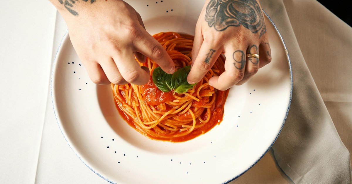 Tattooed hands adding basil to spaghetti with tomato sauce, offering a culinary touch.