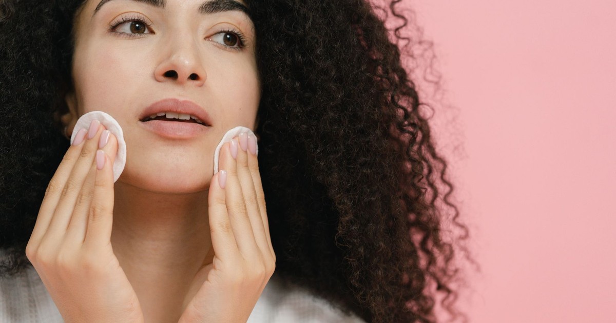Close-up of a woman cleansing her face with cotton pads against a pink background.