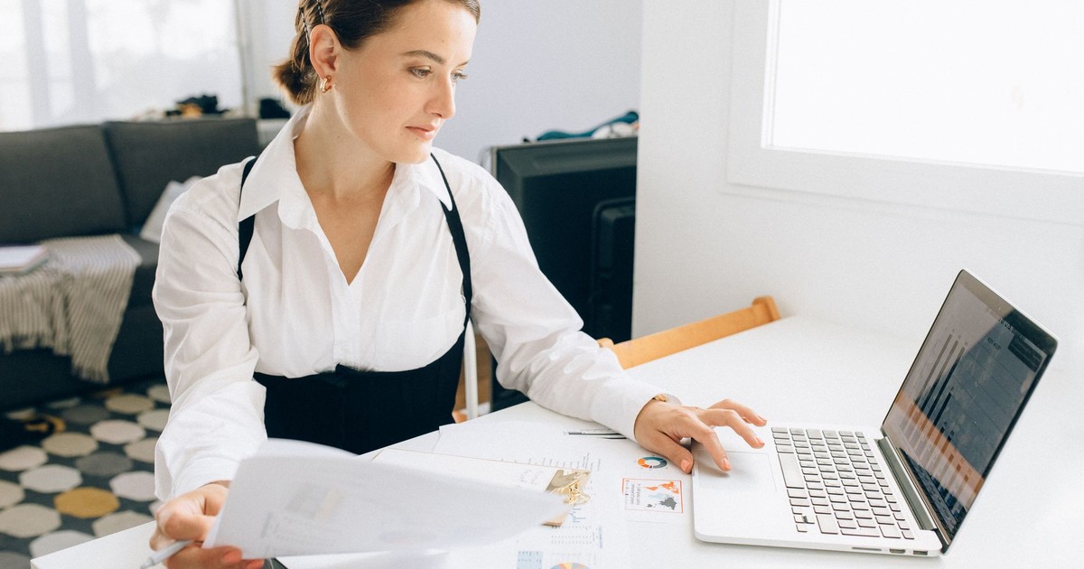 Woman at desk reviewing business reports on laptop, holding papers in a modern office setting.
