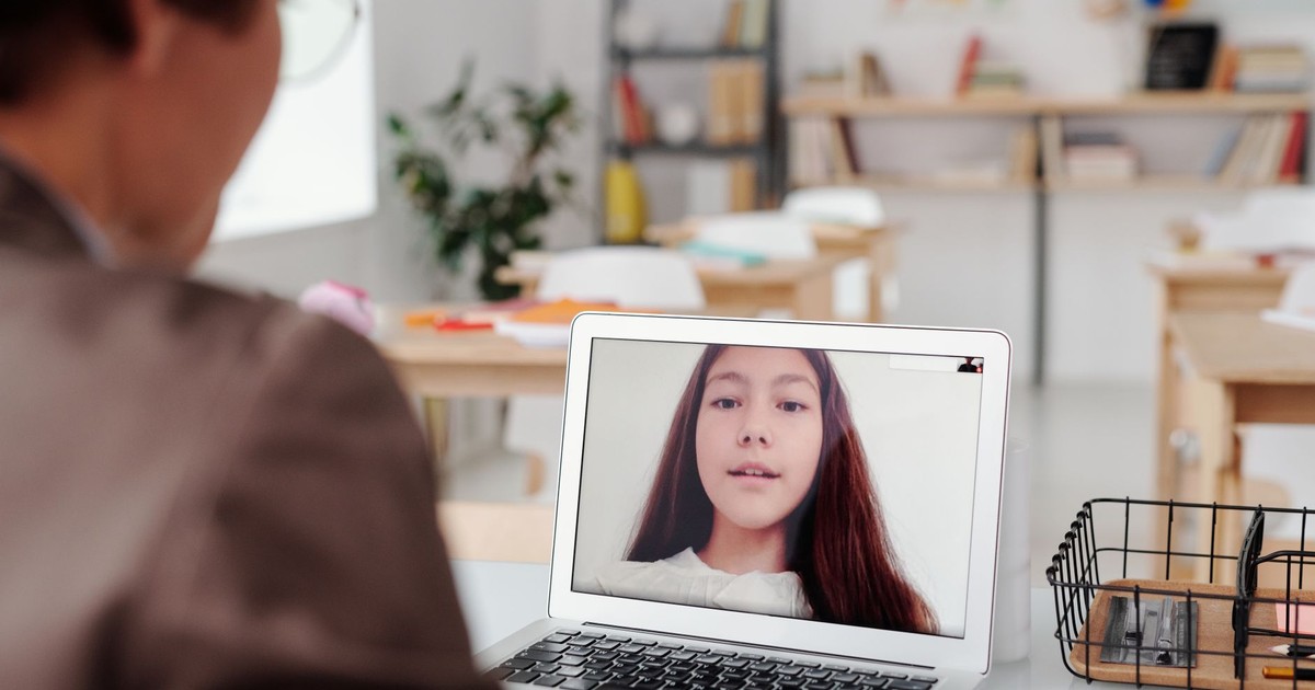 Teacher using laptop for virtual lesson with female student on screen in classroom setting.
