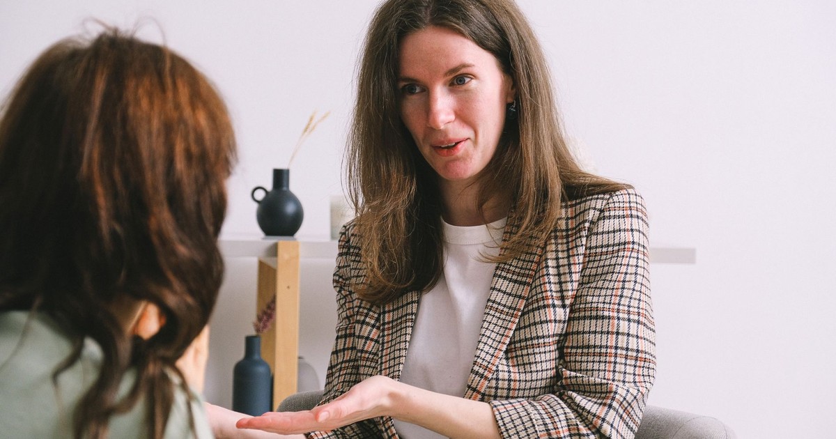 Positive adult female psychologist in smart casual outfit smiling while talking about treatment with unrecognizable woman sitting in comfortable armchair in modern workplace
