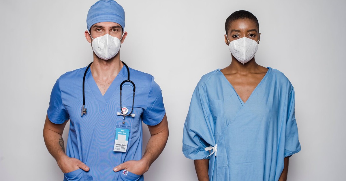 Multiethnic adult male doctor with stethoscope standing near female patient in blue uniform and protective masks in light studio on white background while looking at camera with hands in pockets
