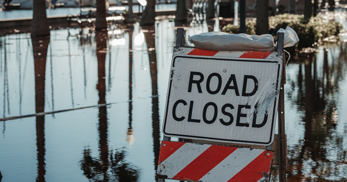 Road closed sign amidst flooded street, reflecting calm water and trees.