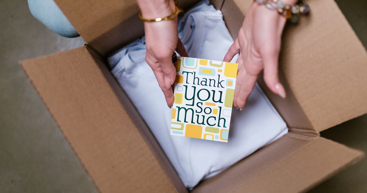 A person holds a thank you card above items in an open shipping box, suggesting care in sending.