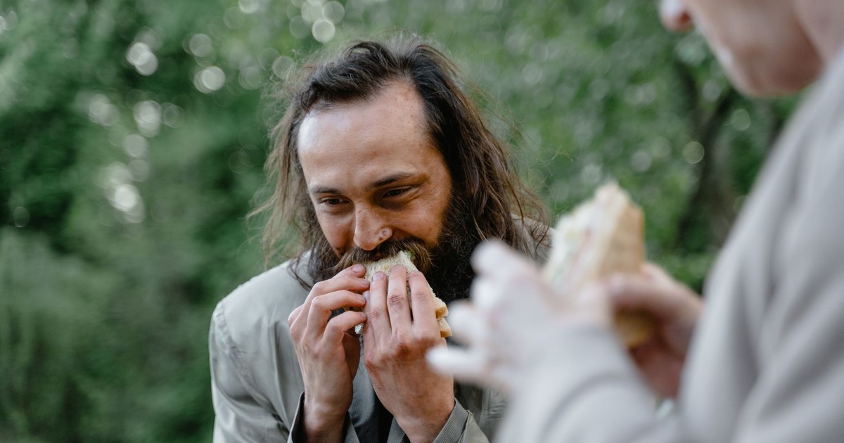 A man happily eats a sandwich outside in a green and natural setting.