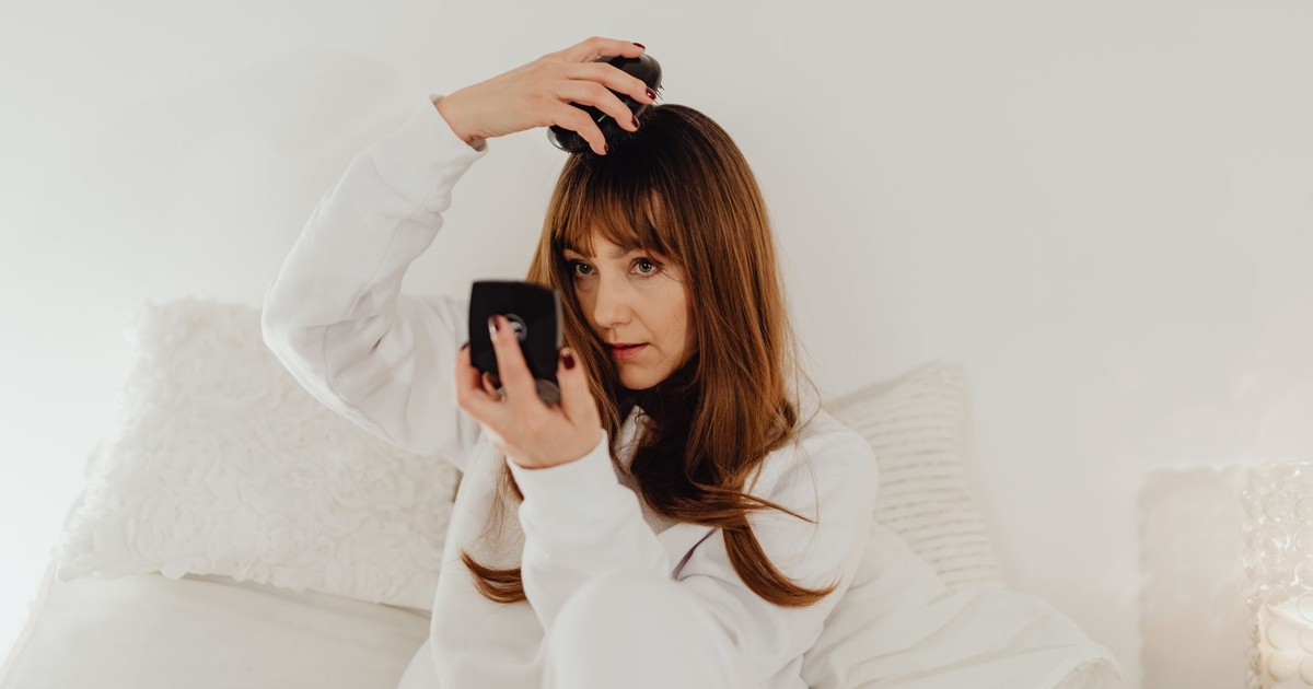 Woman in white attire combs her hair while seated on a bed with soft lighting.