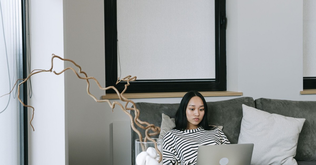 Asian woman working from home on laptop, sitting on a sofa in a modern, minimalist space.