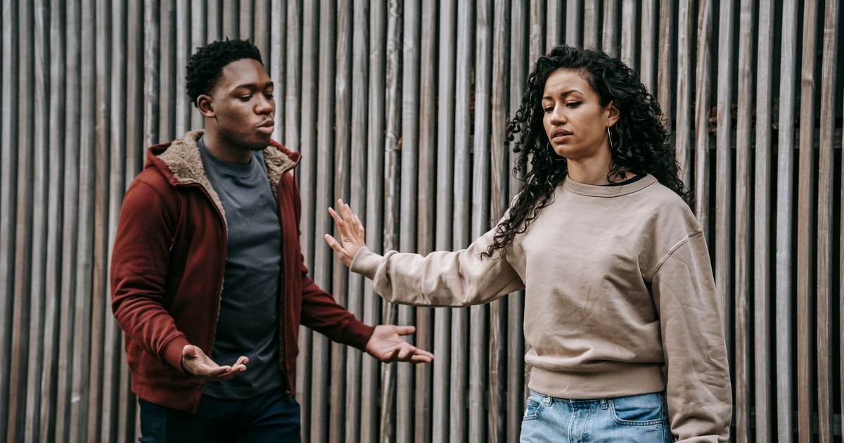 Insulted ethnic girlfriend with outstretched arm and irritated African American boyfriend having quarrel near wooden wall on street during breakup