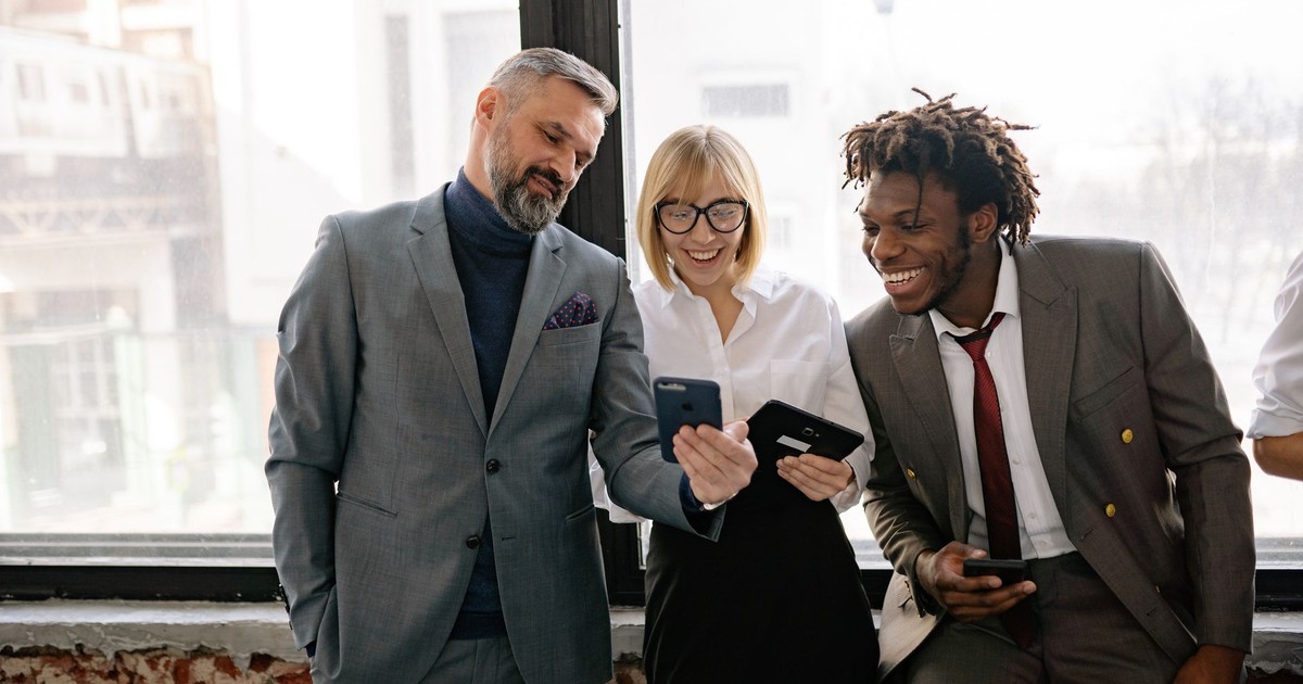 Three colleagues in a modern office sharing a moment over their smartphones.