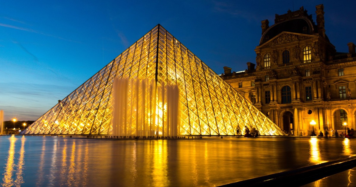The Louvre Pyramid illuminated beautifully in the evening, reflecting in the courtyard's water.