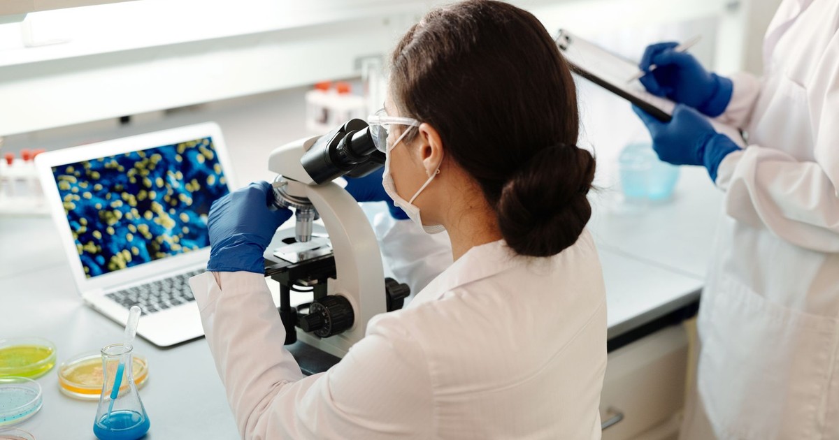 A female scientist in a lab coat examines samples under a microscope for research.