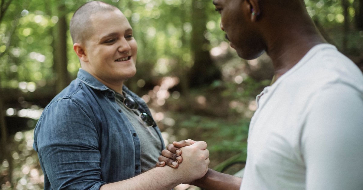 Cheerful young male in jeans shirt standing in green woods and shaking hands with anonymous African American male friend