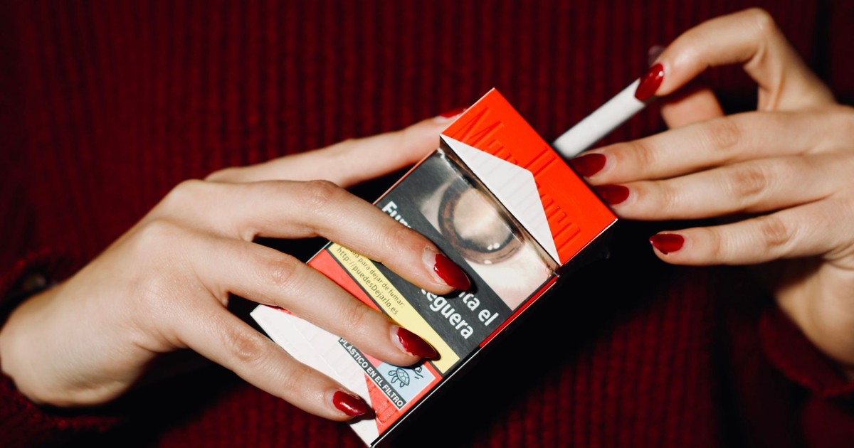 Close-up of hands with red nails holding a cigarette pack indoors.