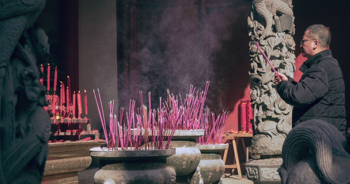 A man lights incense sticks in an Asian temple, surrounded by intricate sculptures.