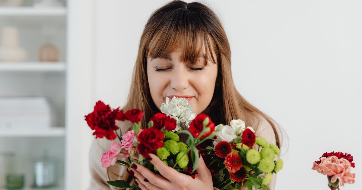 A woman indoors holding a bouquet of colorful flowers, enjoying the fragrance with eyes closed.