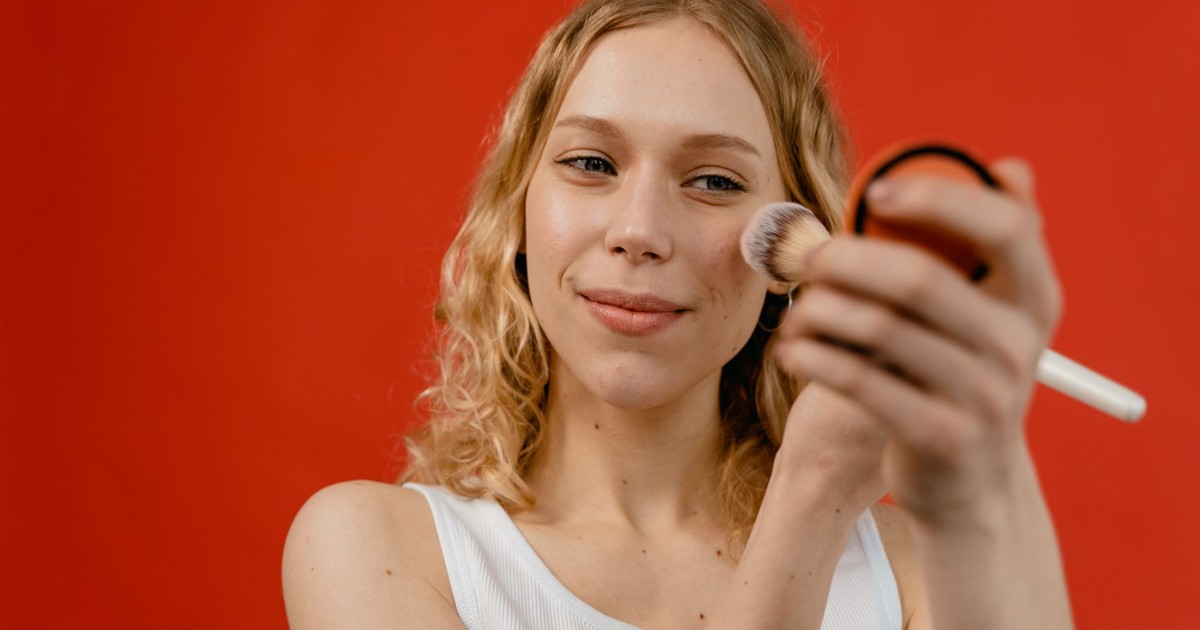 Woman applying makeup with a brush against a vibrant red backdrop, showcasing beauty and skincare concepts.