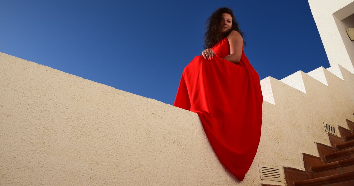 Woman in red dress posing elegantly against blue sky and white architecture.