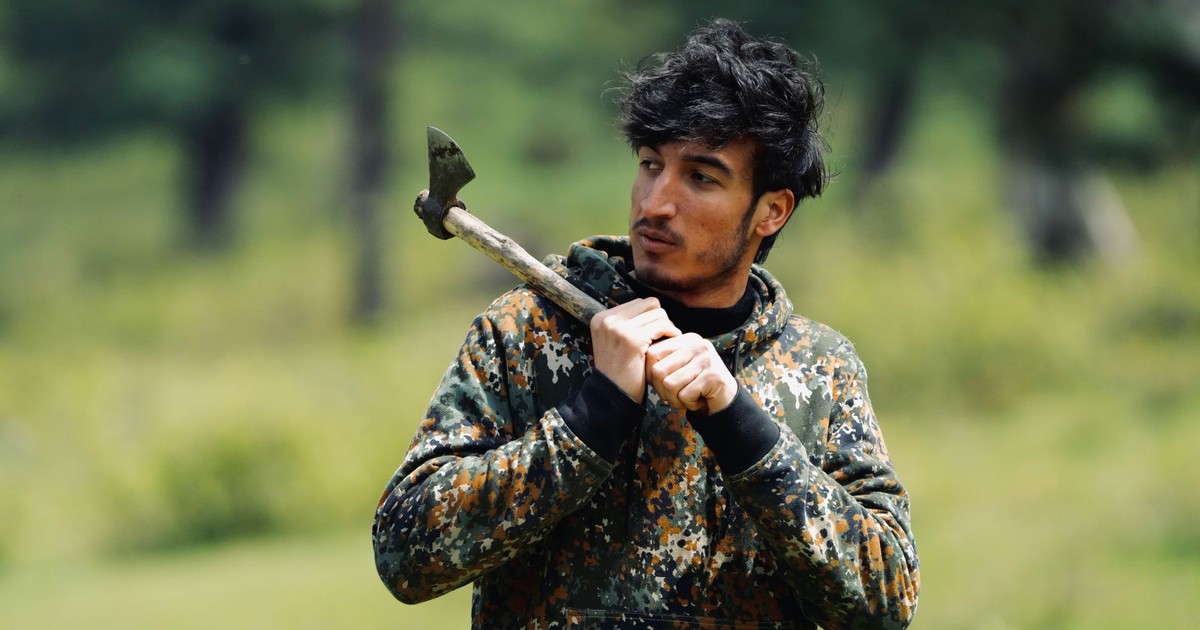 A young man in a camouflage hoodie holding an axe while standing outside in a forest park.