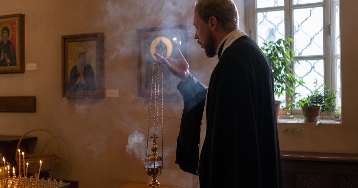 Orthodox priest holding censer in church, surrounded by religious icons and smoke.