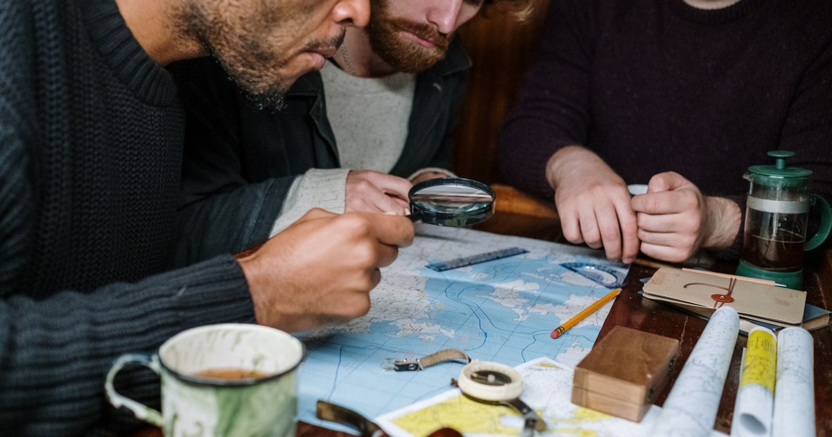 Three men are intensely discussing plans using maps and a magnifying glass, creating a scene of collaboration.