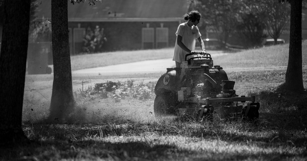 A person operating a lawnmower in a shaded, grassy yard, in black and white.
