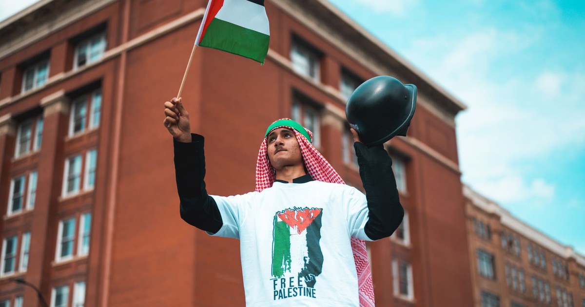 Man waving a Palestine flag in protest with an urban backdrop.