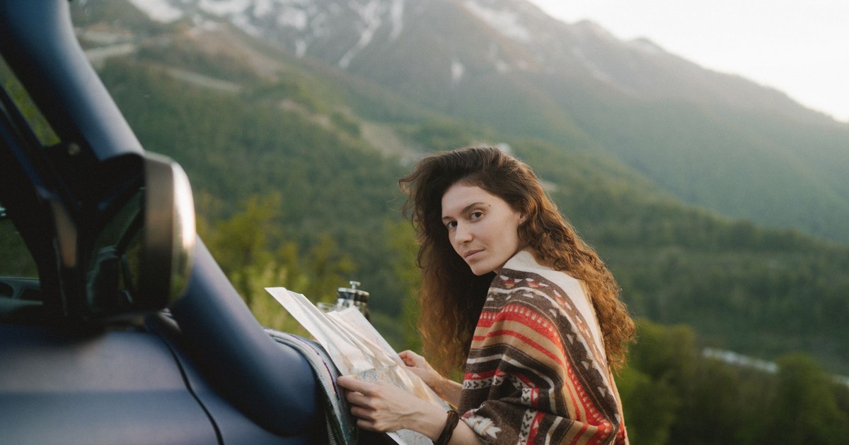 A woman with long hair holding a map by a car in a scenic mountainous landscape.