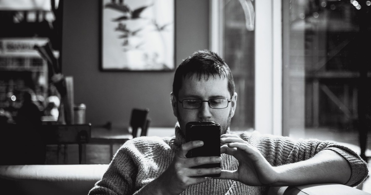 A man in a cozy indoor space engrossed with his smartphone, sipping coffee.