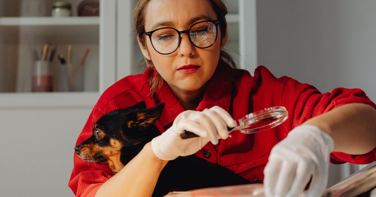 A woman wearing glasses examines documents with a magnifying glass, holding her dog.