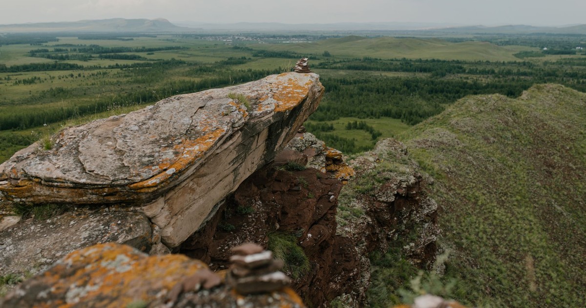Drone view of stony rough terrain with uneven surface near green meadows with herb and forests under cloudy sky